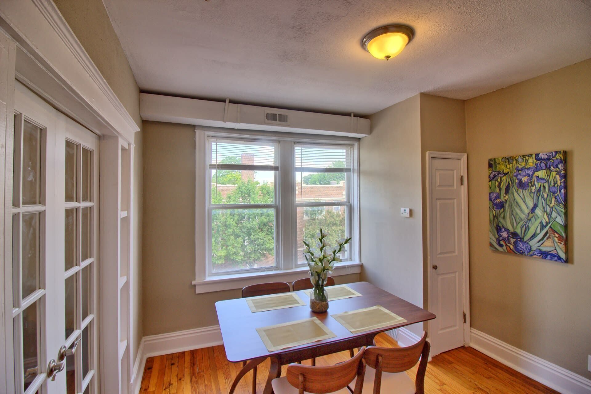 A dining area with a table, chairs, a vase of flowers, a painting on the wall, and a door.