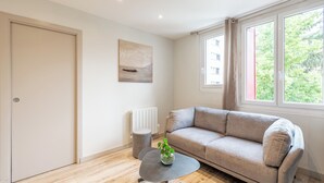 Een moderne woonkamer met een beige bank, een klein rond tafeltje met een plant, een radiator en een raam met uitzicht op gebouwen en bomen.