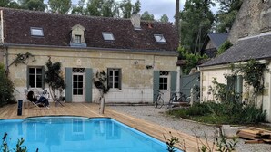 En swimmingpool med ett trädäck, en byggnad i sten med gröna fönsterluckor och en cykel parkerad utanför.