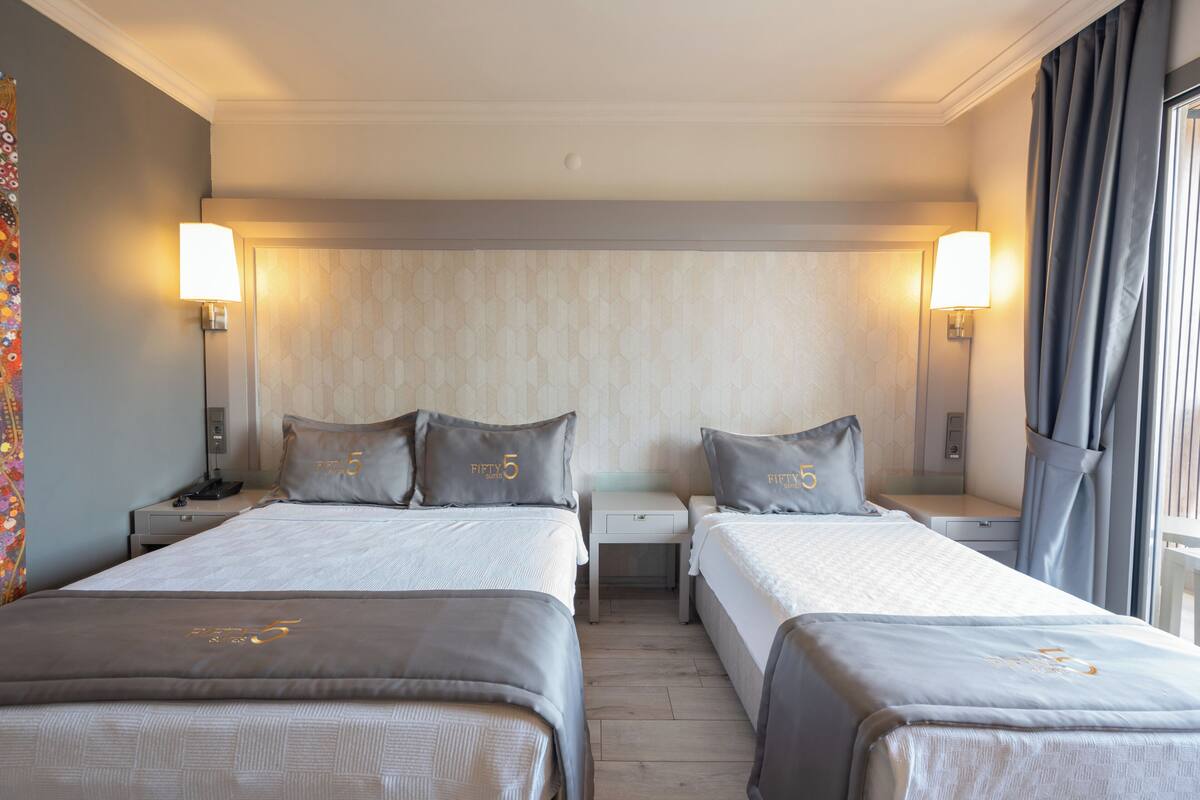 A hotel room with two beds, a nightstand with a lamp, a small table, and a window with curtains.