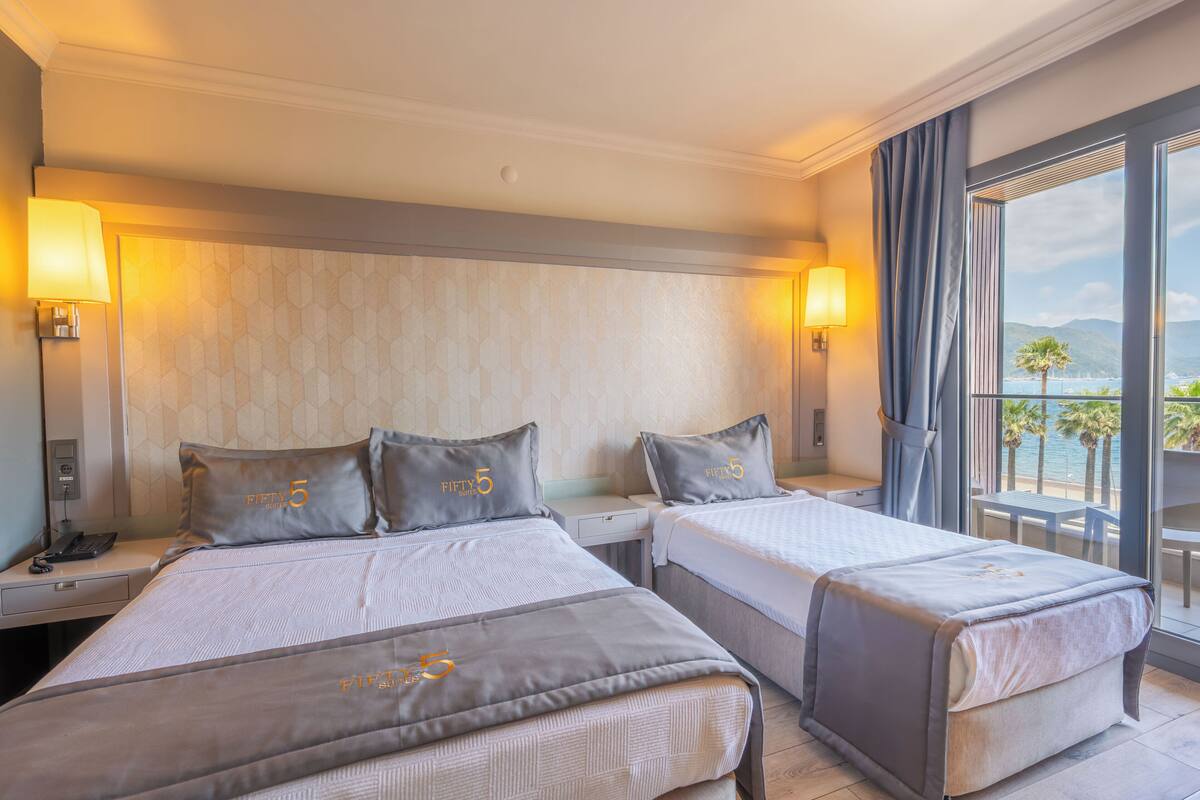 A hotel room with two beds, a large window with a view of the sea, and a balcony with outdoor seating.