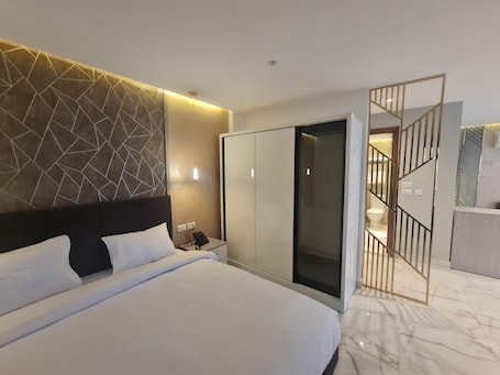 A modern hotel room with a large bed, a wardrobe, a desk with a phone, and a bathroom visible through a glass door.