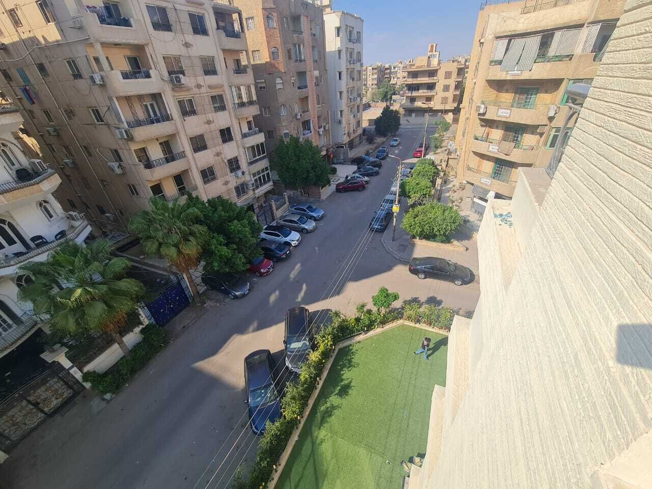 A residential area with multi-story buildings, parked cars, and a green lawn.