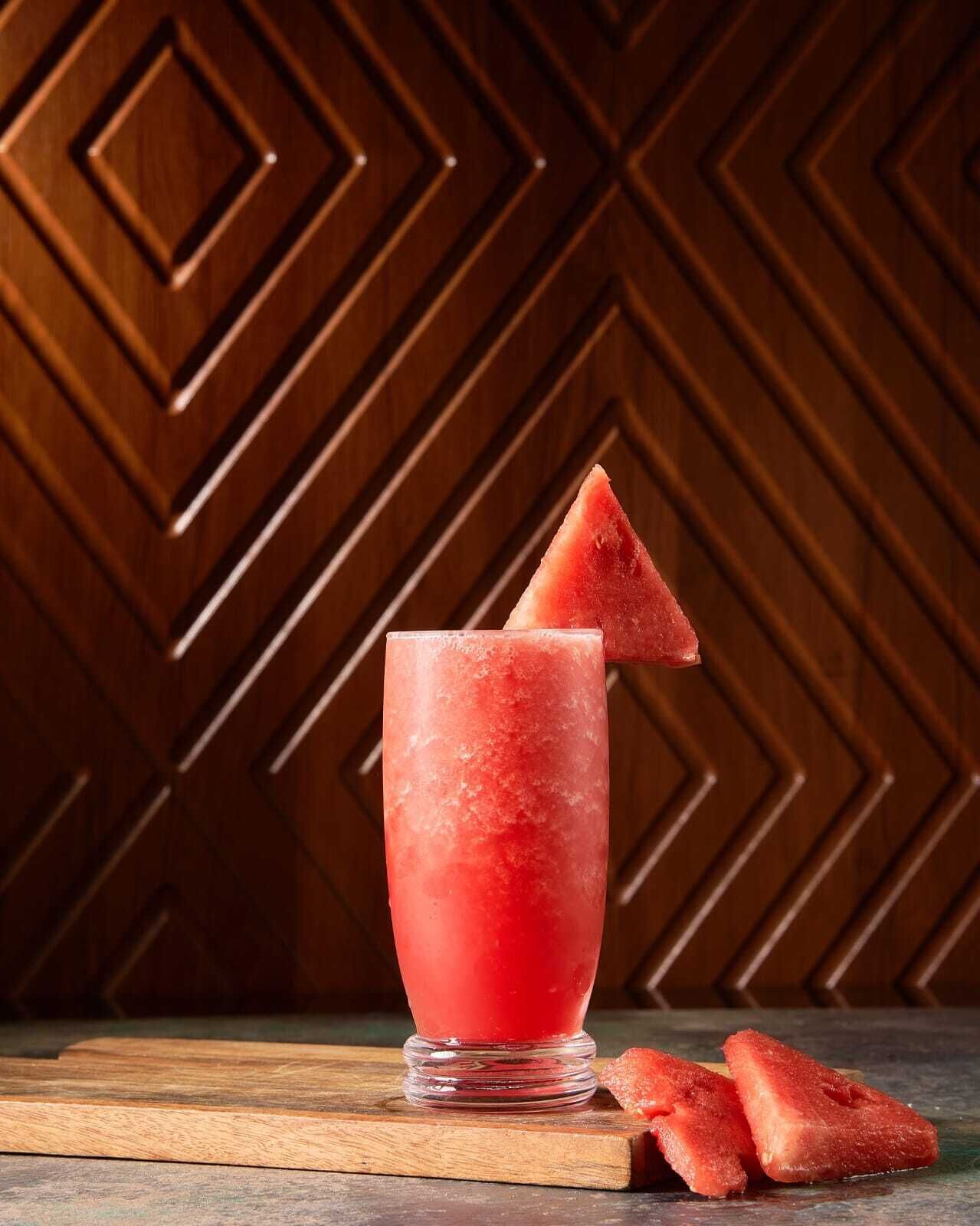 Un verre contenant une boisson rouge, orné d’une tranche de pastèque sur le bord, posé sur une surface en bois avec un fond à motifs.