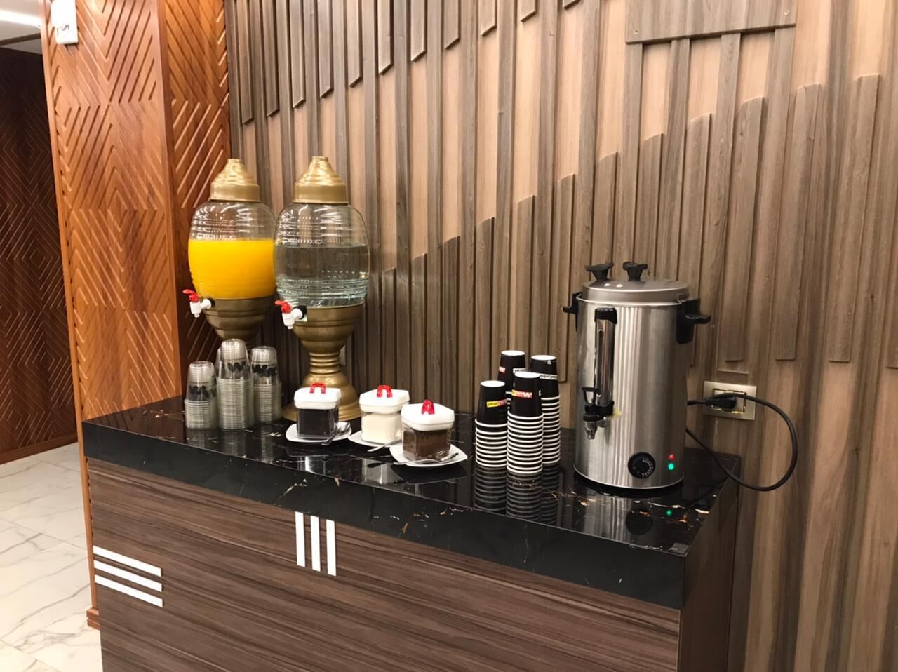 Zona de recepción de un hotel con un mostrador de mármol negro, una m áquina de café y varios vasos y recipientes para café.