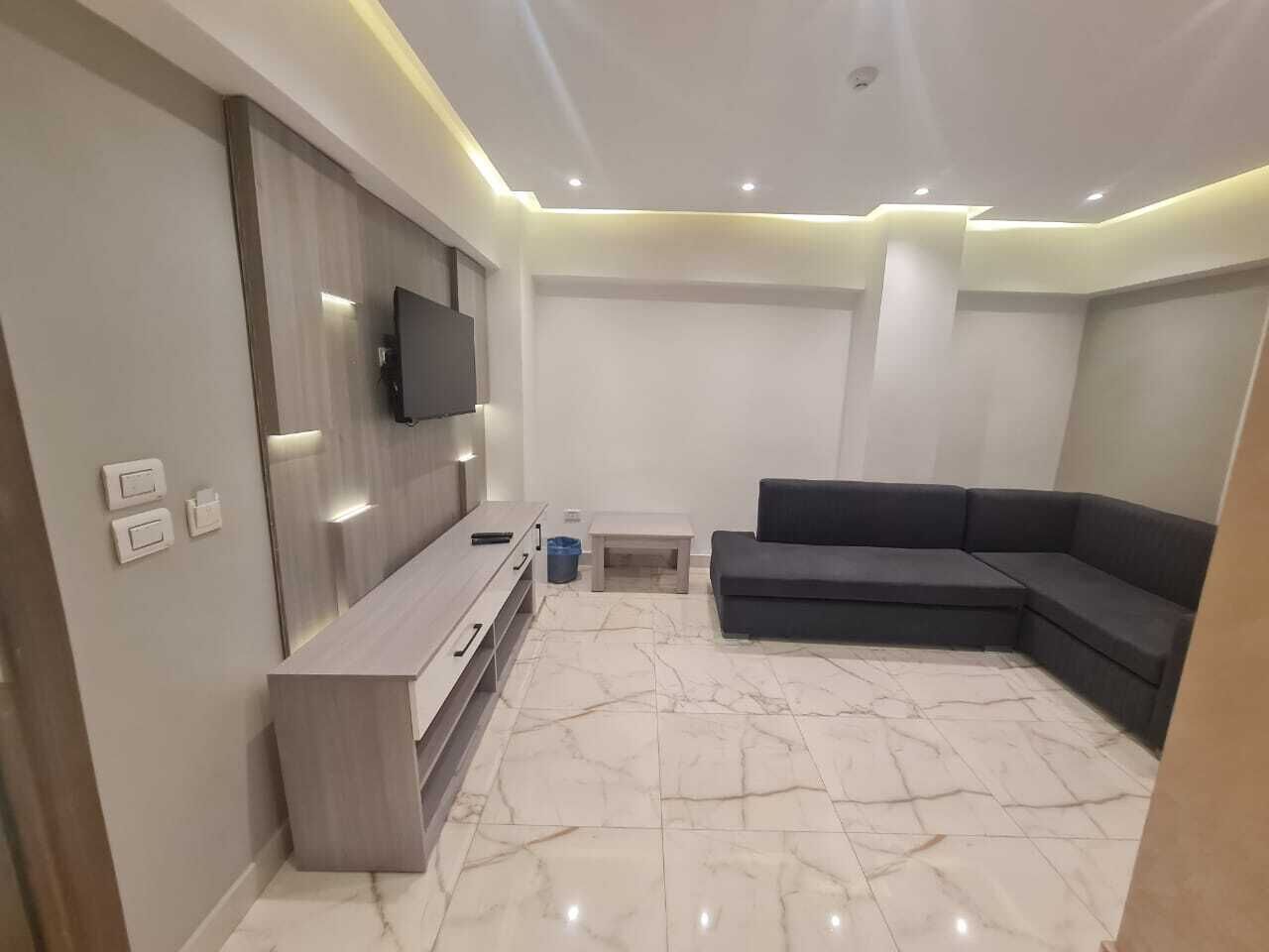 A modern living room with a flat-screen TV mounted on the wall, a grey sectional sofa, a wooden coffee table, and a marble-patterned floor.