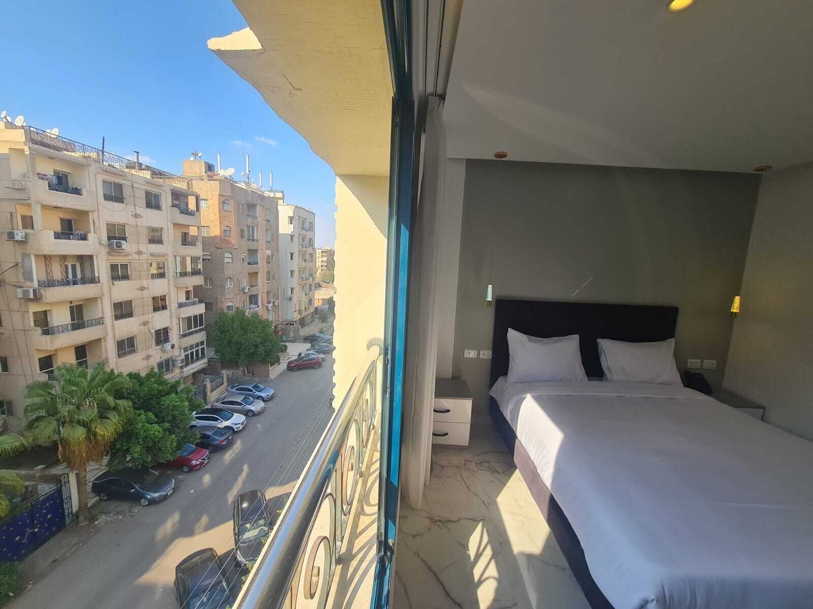 Ein Balkon mit einem Bett, Blick auf eine Straße mit geparkten Autos und mehrstöckigen Gebäuden im Hintergrund.