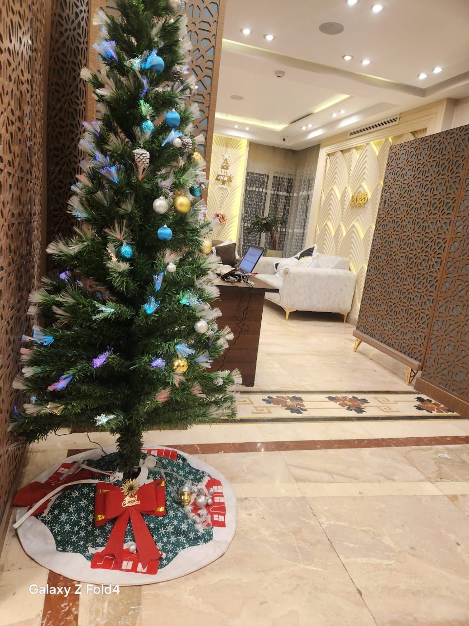 A Christmas tree with ornaments, a laptop on a desk, a sofa, and a patterned wall.