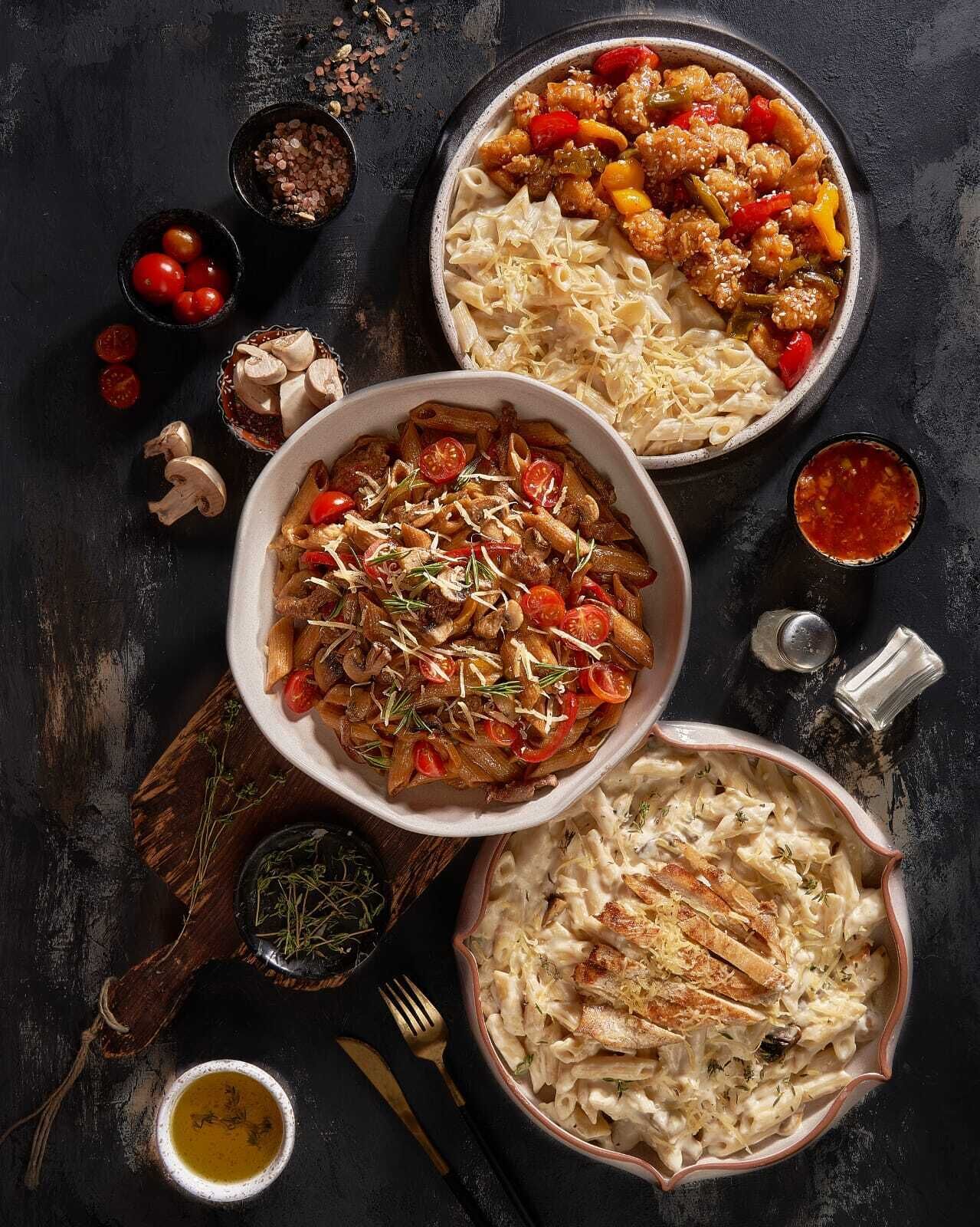 Three bowls of pasta dishes with various toppings and garnishes, accompanied by scattered cherry tomatoes, mushrooms, and herbs on a dark surface.