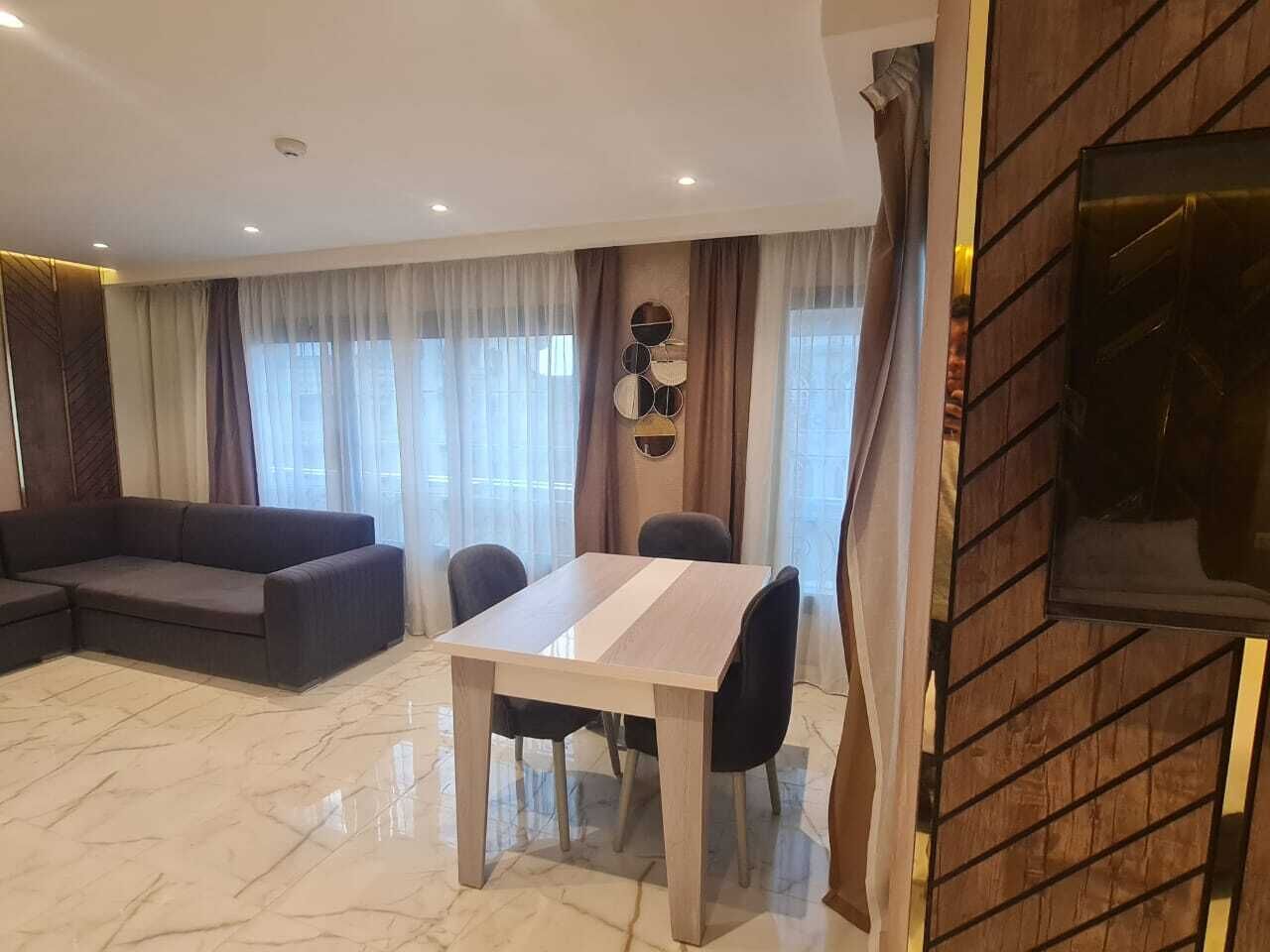 A modern living room with a sofa, dining table, and chairs. There is a TV mounted on the wall and a wooden panel on one side.