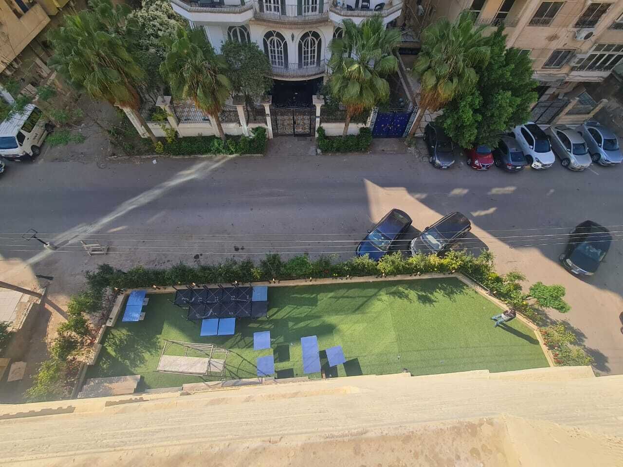 A courtyard with a green lawn, a few parked cars, and a building with a balcony in the background.
