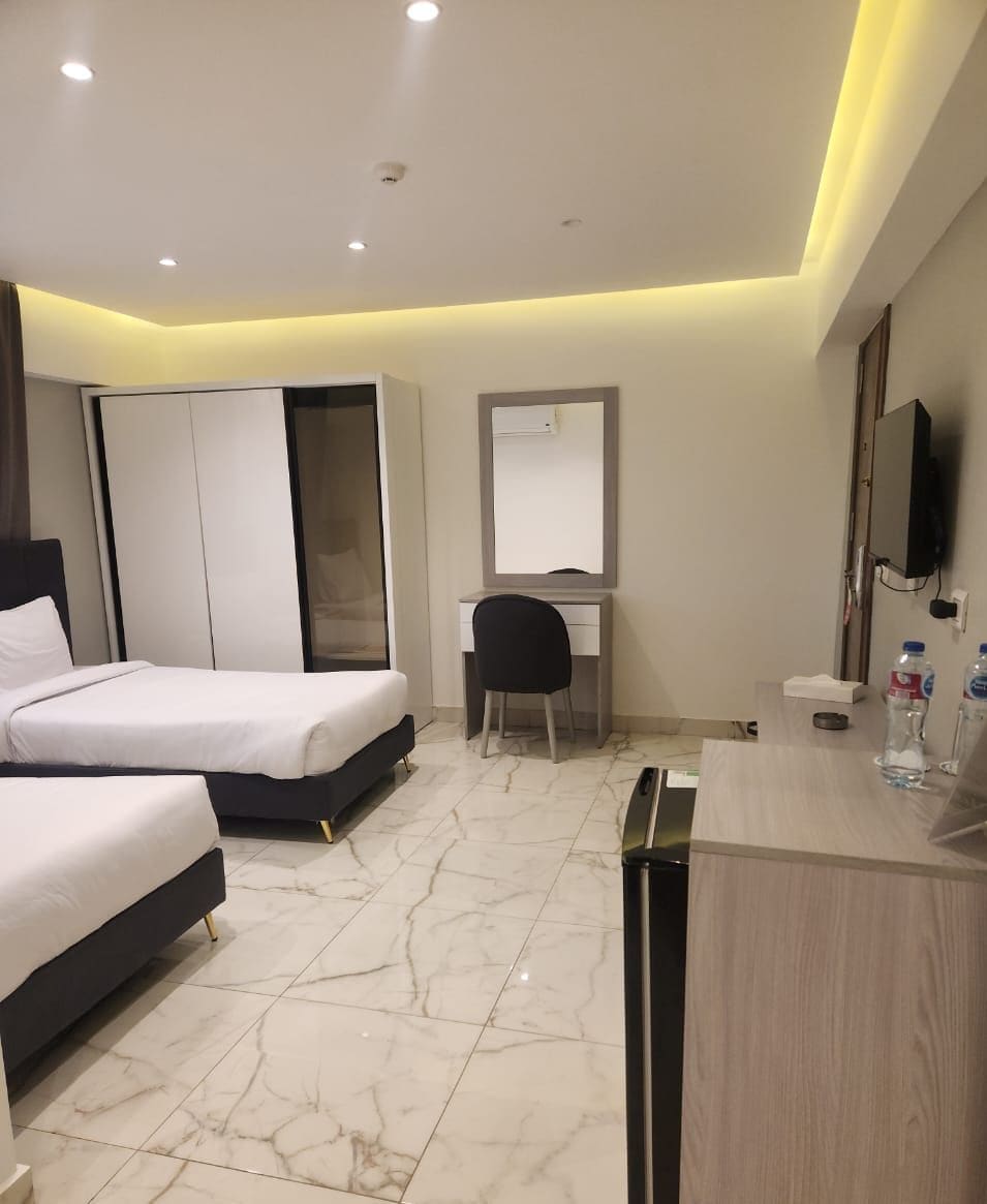 A hotel room with two beds, a desk, a chair, a mirror, a TV, and a bathroom area.