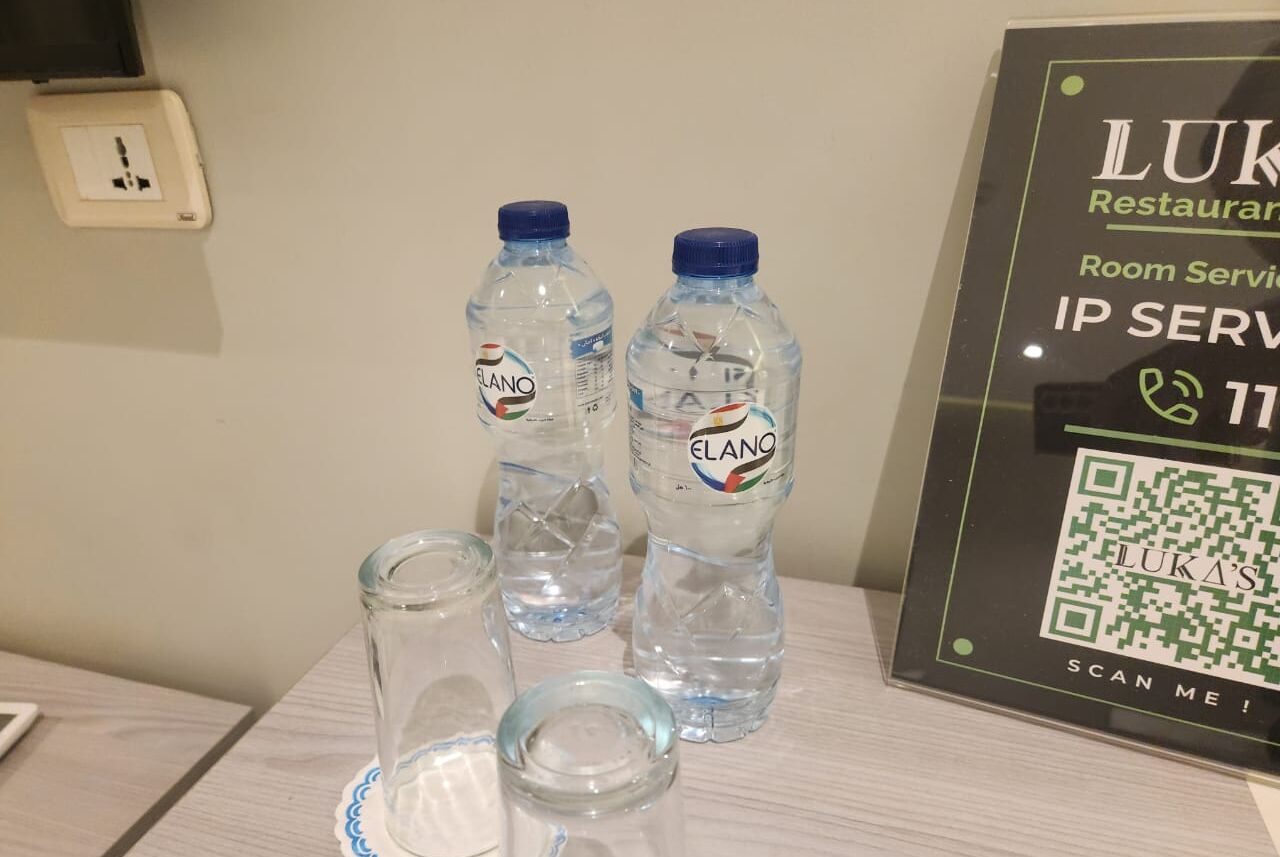 Una mesa con dos botellas de agua etiquetadas 'ELANO' y un vaso. Se ve un código QR y texto que indica 'Restaurante LUKA' y 'Servicio a la habitación' en la pared.