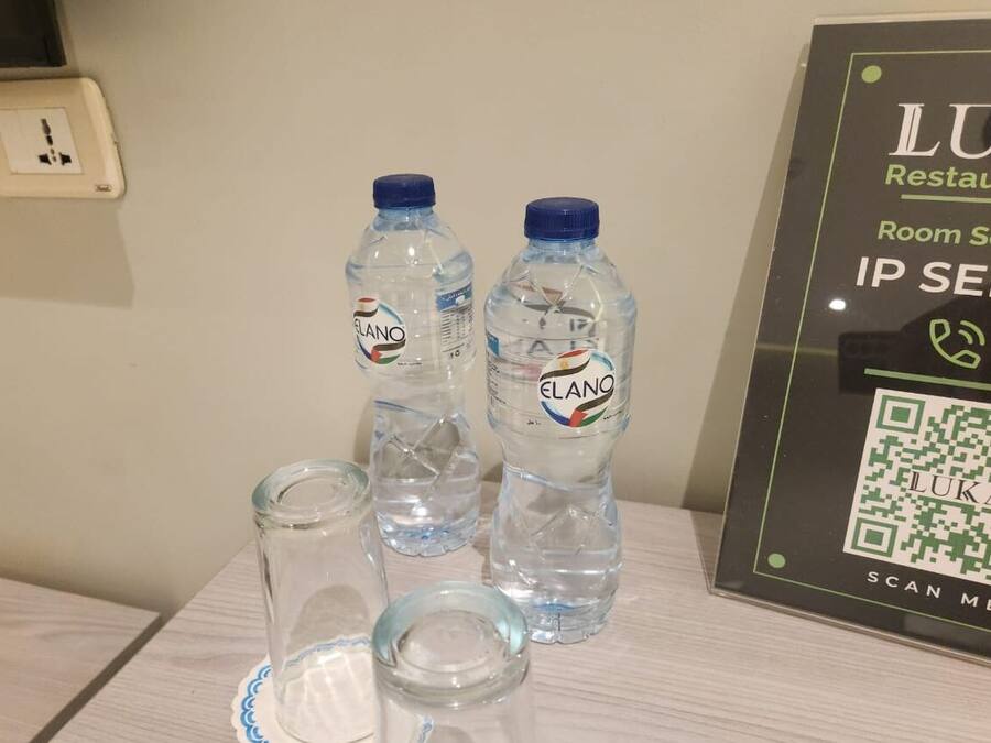 Una mesa con dos botellas de agua etiquetadas 'ELANO' y un vaso. Se ve un código QR y texto que indica 'Restaurante LUKA' y 'Servicio a la habitación' en la pared.