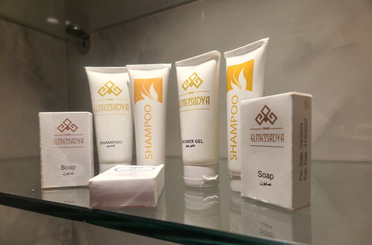 A bathroom shelf with various toiletries including a bar of soap, two tubes of shampoo, and a bottle of shower gel, all branded with 'ELMESSADYA'.