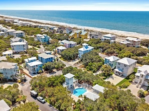 An aerial view of a coastal residential area with houses, a swimming pool, and a beach in the background.