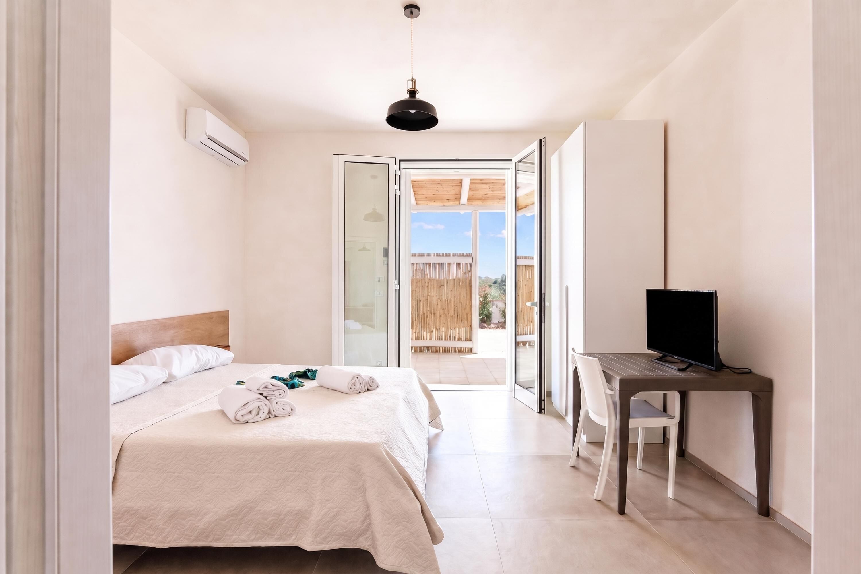A hotel room with a bed, a desk with a television, and a sliding glass door leading to an outdoor area.