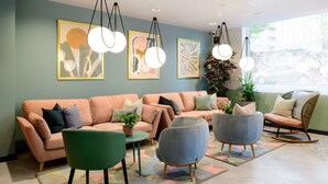 A modern living room with a peach-colored sofa, green and gray armchairs, and a rocking chair. There are abstract paintings on the wall and a potted plant on the floor.