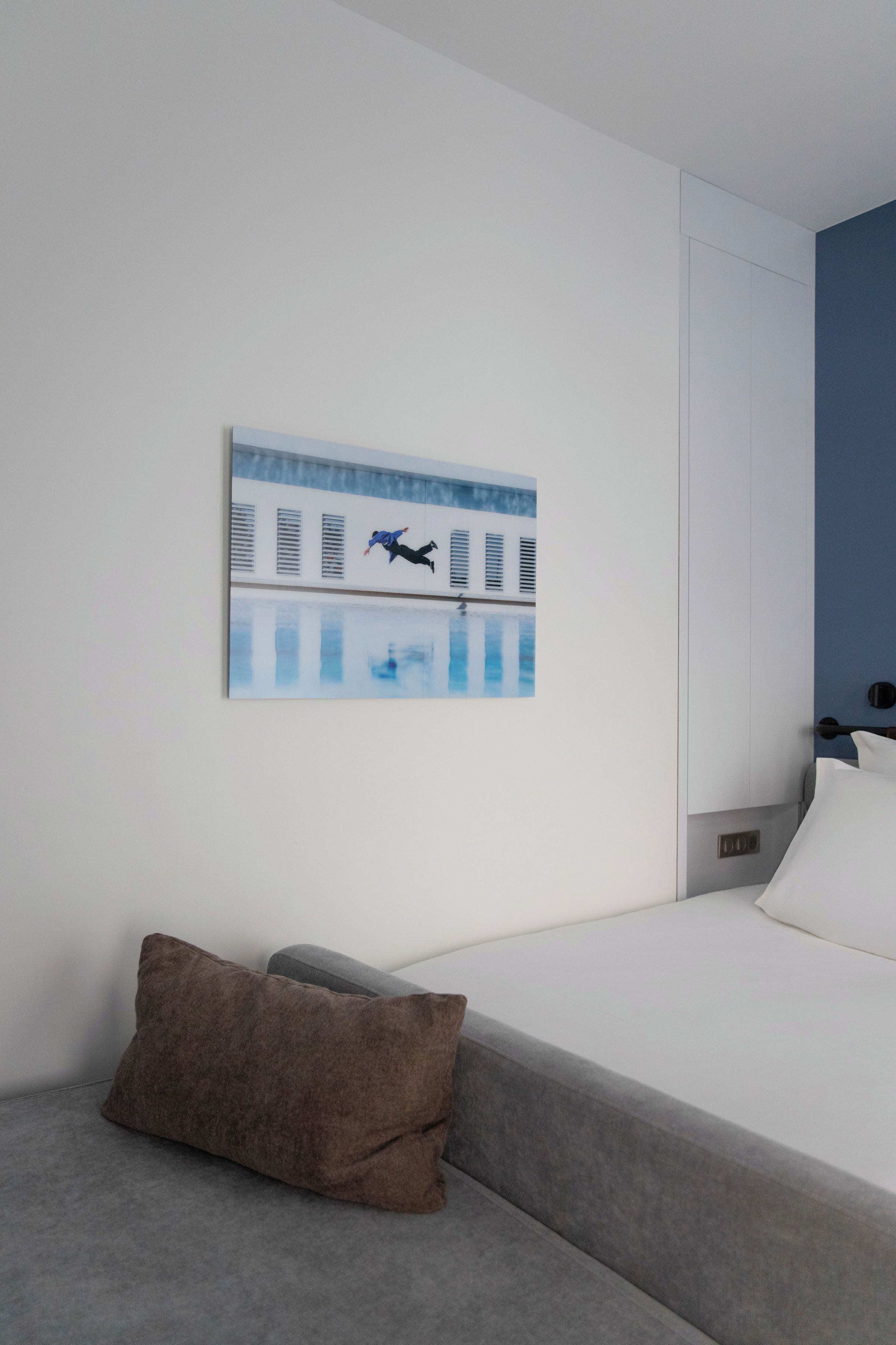 A modern hotel room with a grey sofa, a white bed, and a painting of a person jumping into a pool.