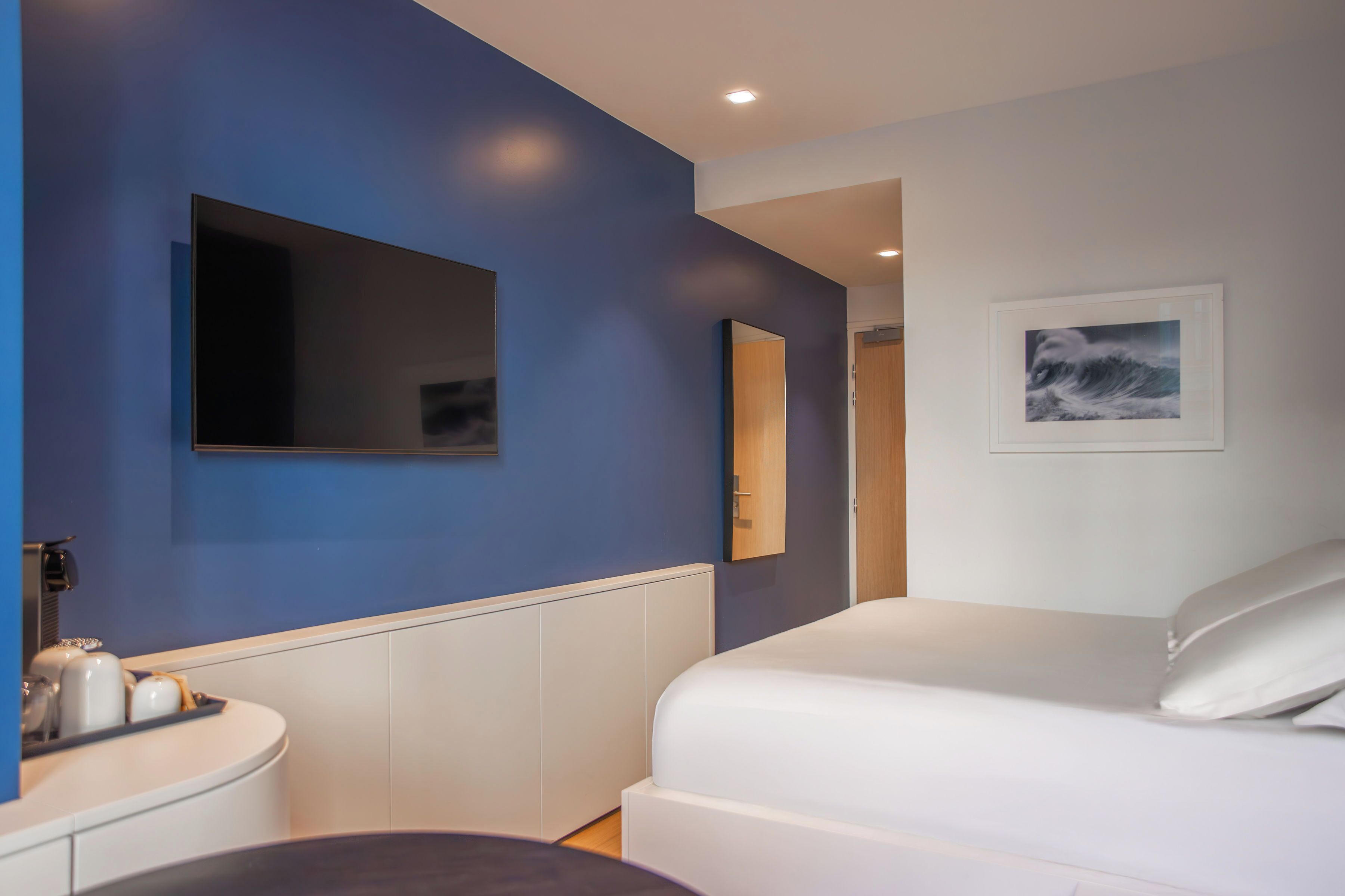 A modern hotel room with a large bed, a flat-screen TV mounted on the wall, a small bedside table, and a framed picture on the wall.