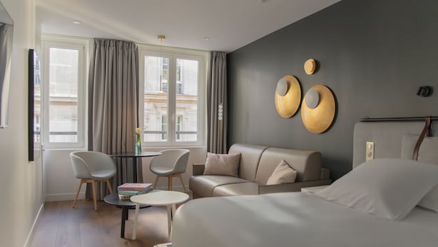 A modern hotel room with a bed, a sofa, two chairs, a small table, and a view of the city through large windows. - Hotel Leev Victoire & Spa (Paris)