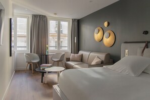 Moderni hotellihuone, jossa on sänky, sohva, kaksi tuolia, pieni pöytä ja suuret ikkunat, joista avautuu näkymä kaupunkiin.