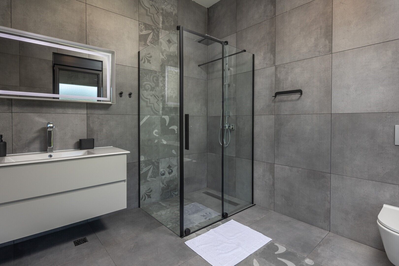 Un baño moderno con ducha acristalada, tocador blanco con espejo y paredes con azulejos grises.