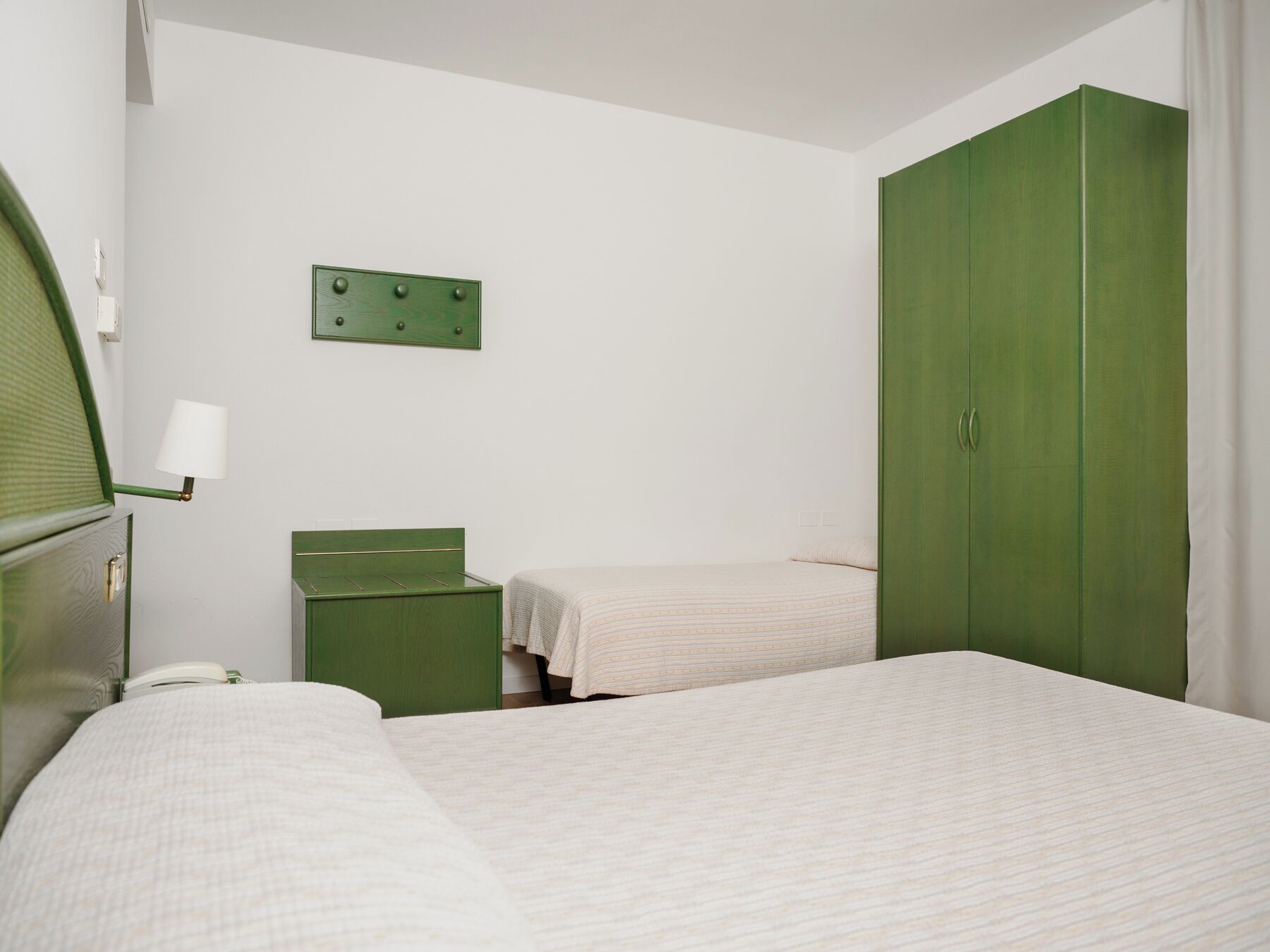 A room with two beds, a green wardrobe, a green nightstand, and a wall-mounted light fixture.
