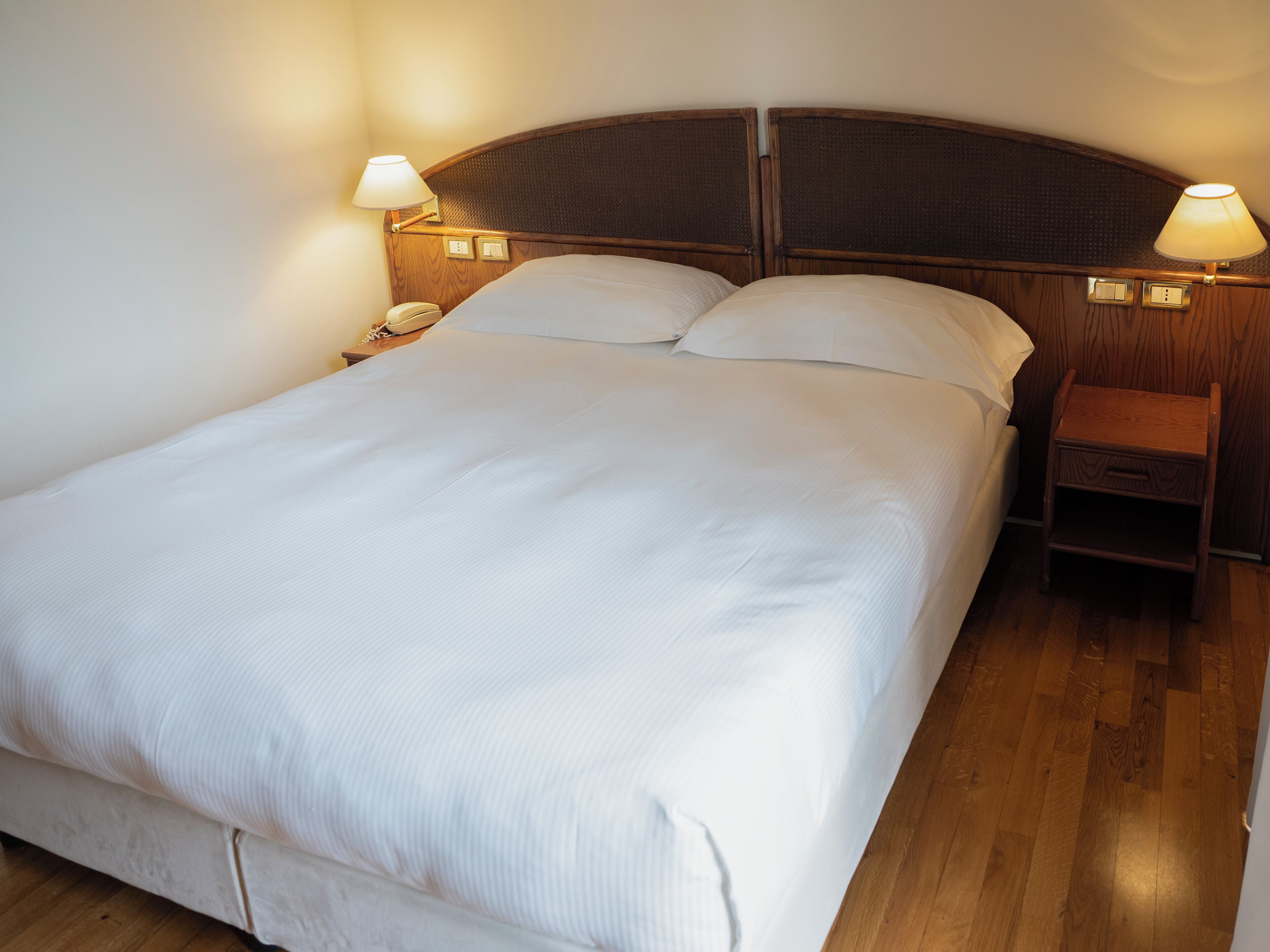 Een net opgemaakt bed met witte lakens, een houten hoofdbord en bedlampjes.