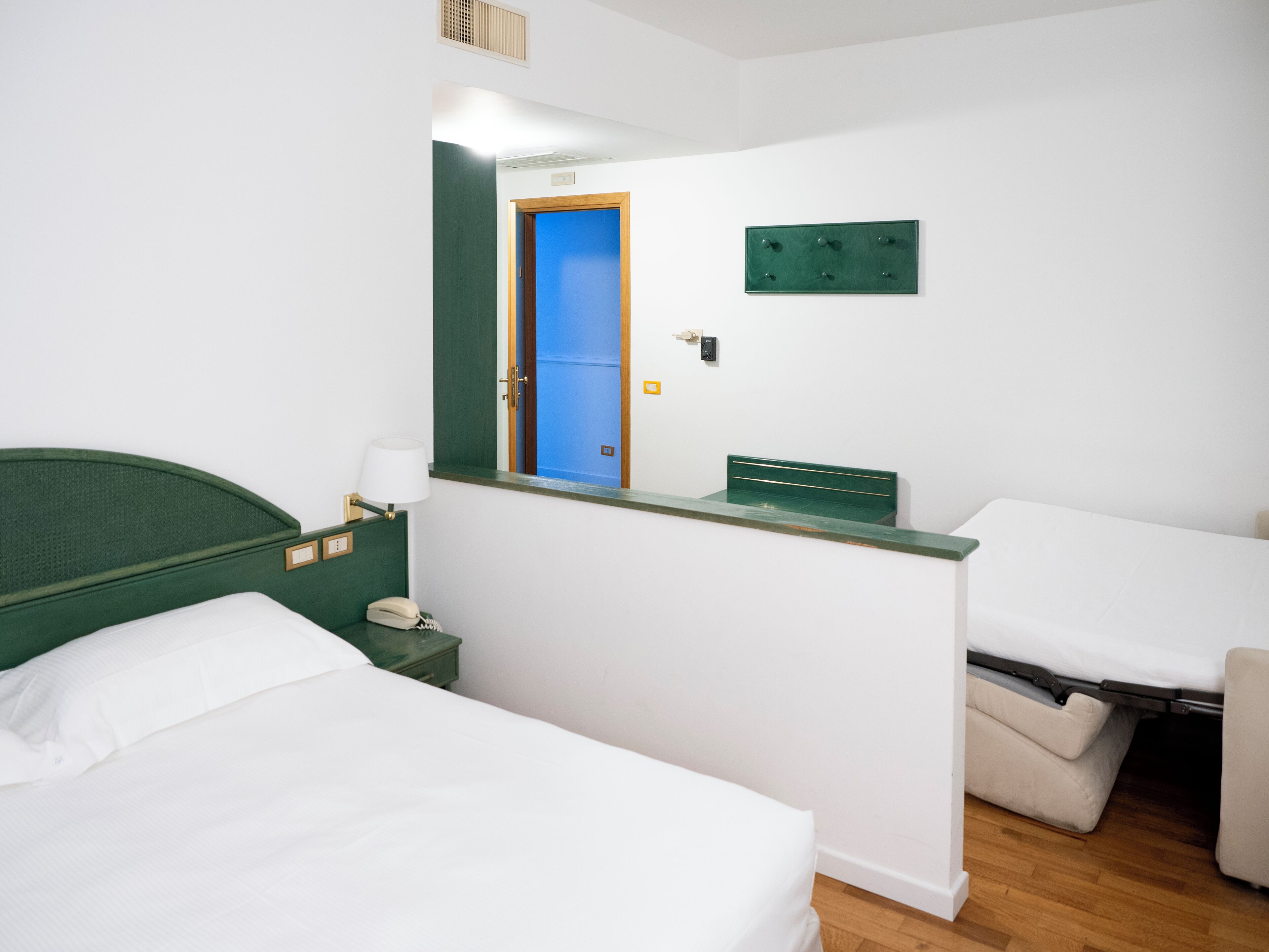 Habitación de hotel con dos camas, un escritorio pequeño con lámpara, cabecero verde, espejo y puerta azul.