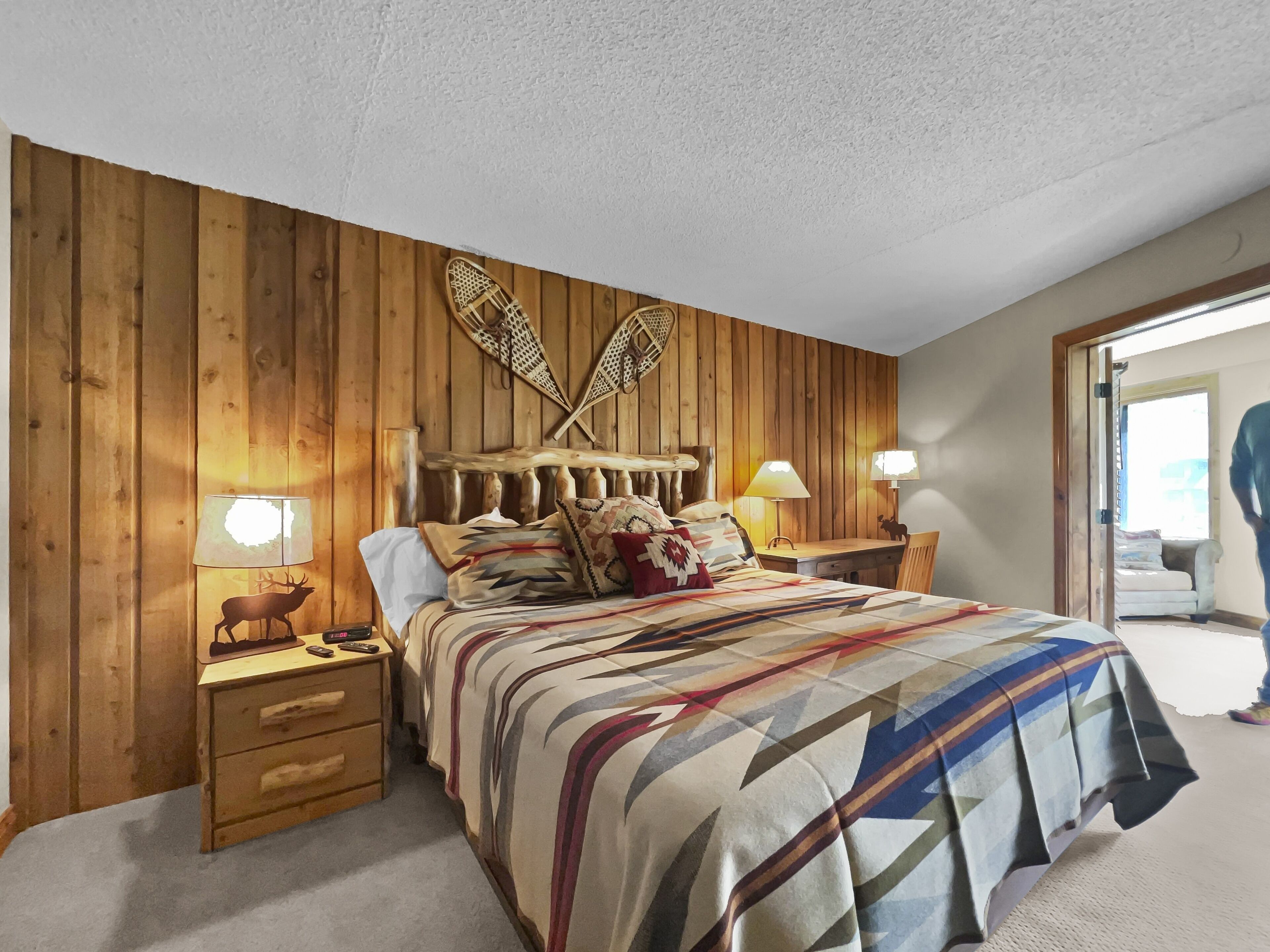 A bedroom with a wooden headboard, a bed with patterned bedding, two bedside lamps, a nightstand with drawers, and a small table with a lamp.