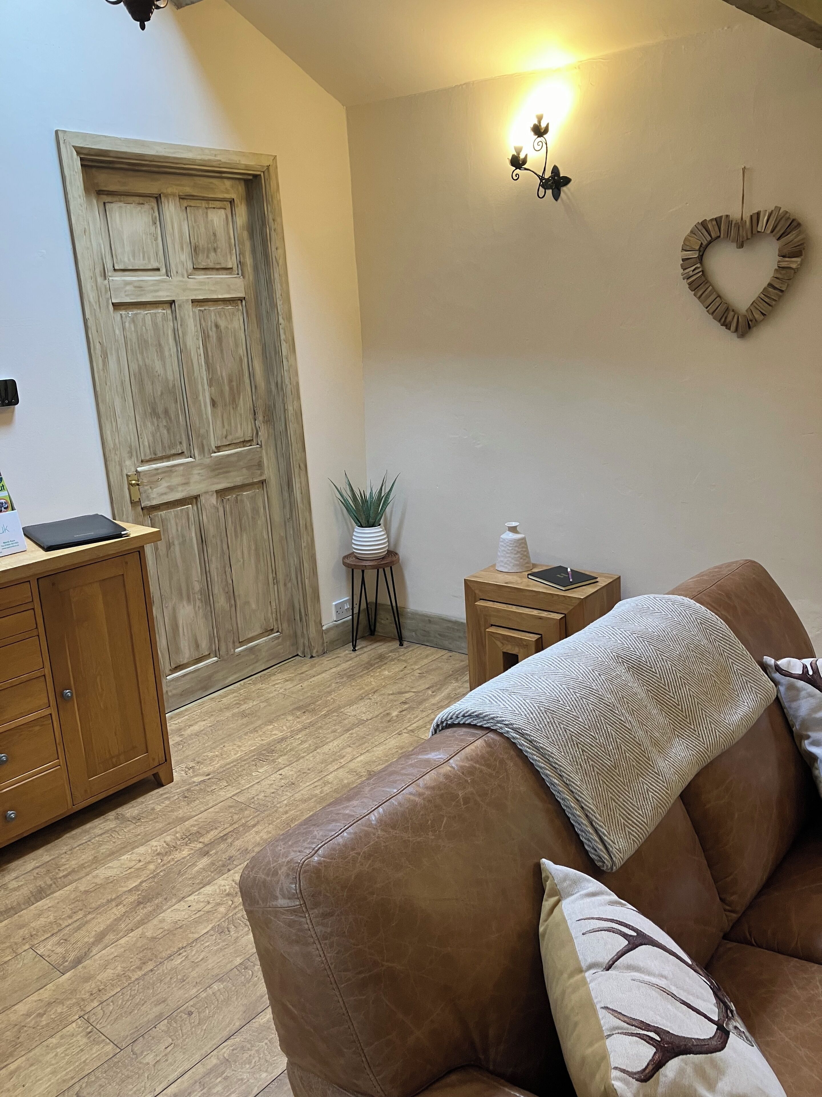 Een gezellige woonkamer met een bruine leren bank, een houten kast, een klein bijzettafeltje en een wandlamp.