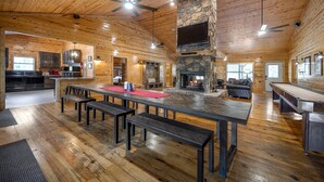 A spacious wooden cabin with a large dining table, benches, a stone fireplace, and a kitchen area.
