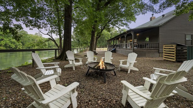A lakeside outdoor seating area with Adirondack chairs arranged around a fire pit, overlooking a body of water.