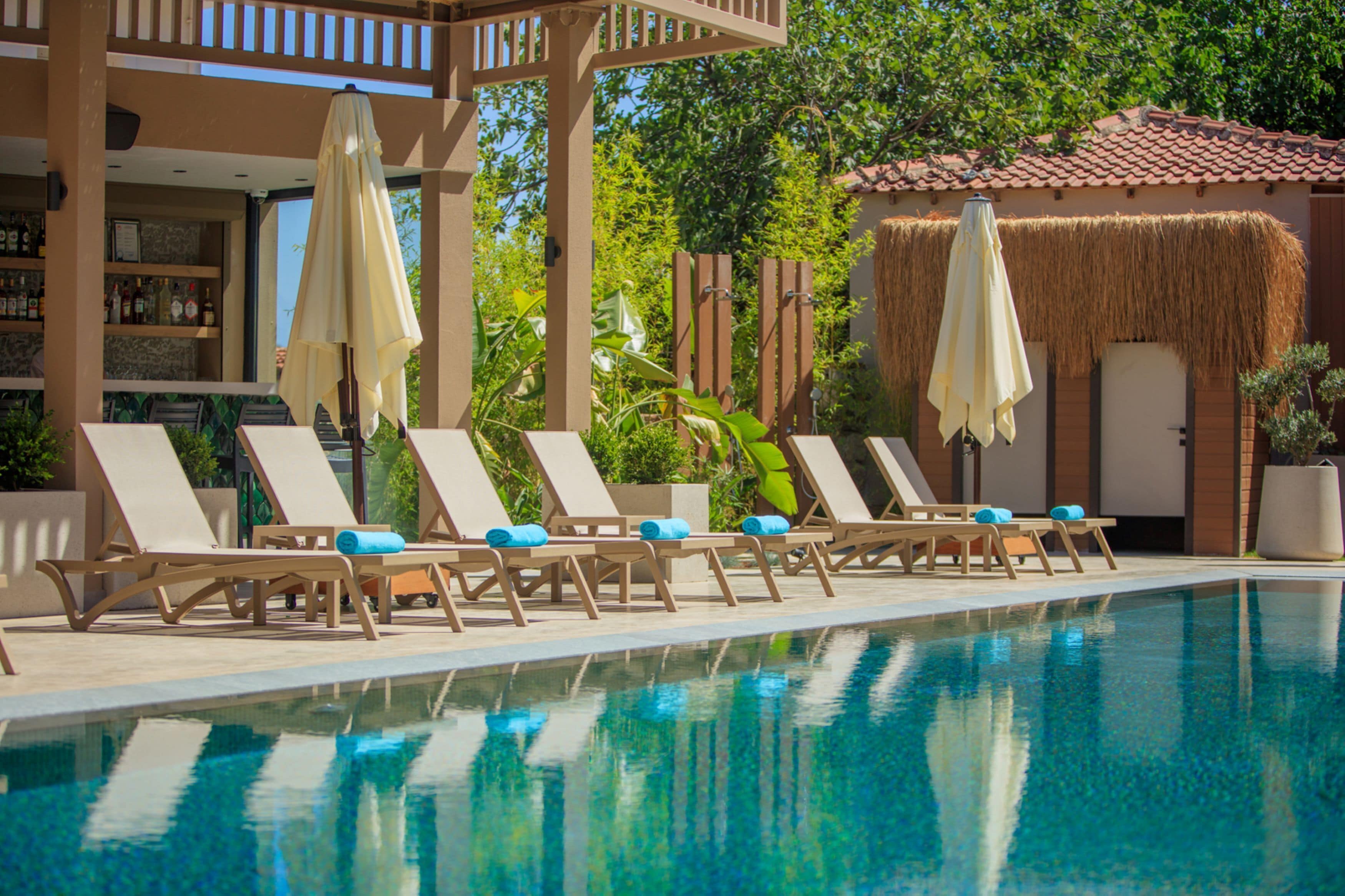 A poolside area with lounge chairs and umbrellas, a bar with shelves of bottles, and a building with a tiled roof.