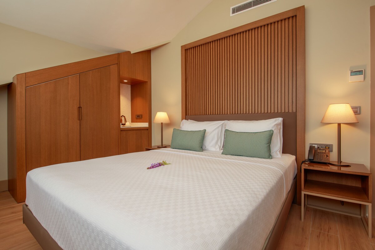 A hotel room with a large bed, wooden wardrobe, bedside lamps, and a small nightstand.