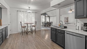 A modern kitchen with dark cabinets, a stainless steel dishwasher, and a dining area with a table and chairs. - Cozy Carolina Trace 3BR w/ Relaxing Golf Course Views - Deck & Fire Table (Sanford)