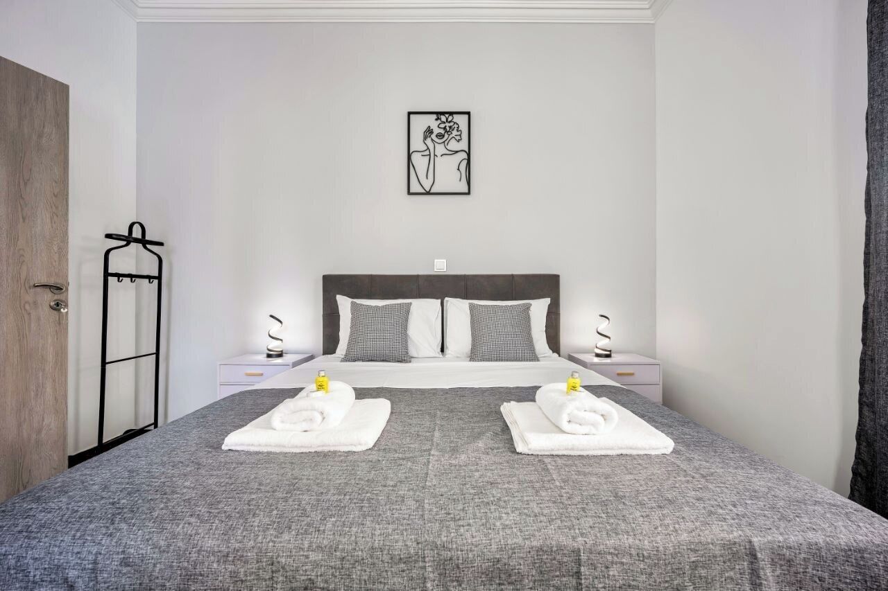 A neatly made bed with two towels and a bottle of lotion on each side, a wall-mounted artwork, and a hanging coat rack.