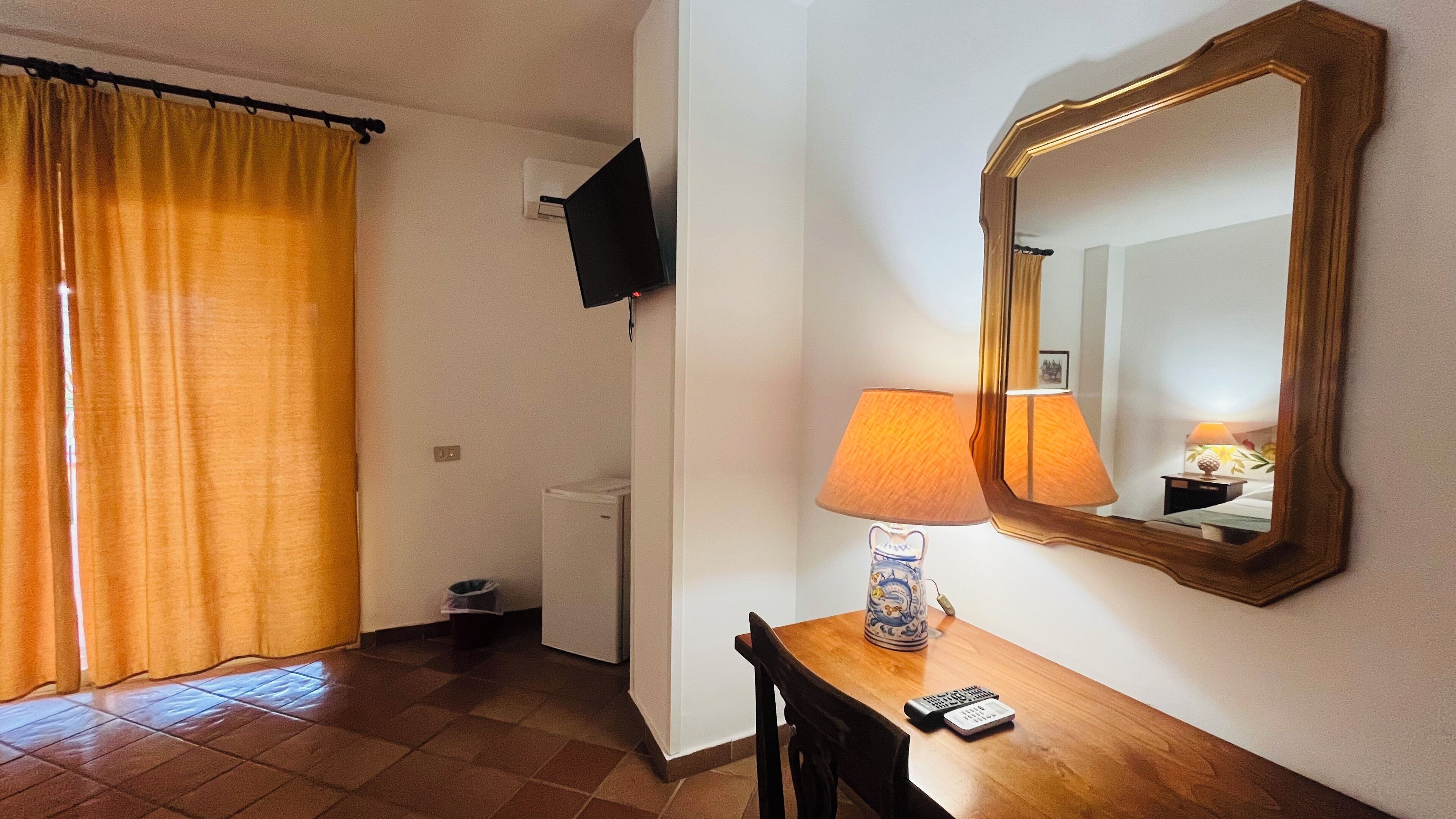 A hotel room with a wooden desk, a lamp, a mirror, a television mounted on the wall, and a window with curtains.