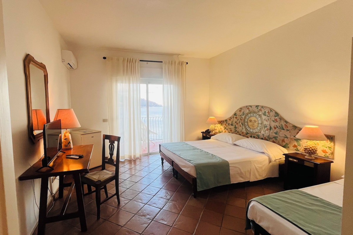 Ein Hotelzimmer mit Bett, Schreibtisch mit Stuhl, Nachttisch und Meerblick durch ein Fenster.