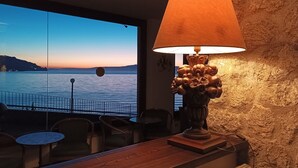 Habitación con vista a un cuerpo de agua al atardecer, una mesa de madera, sillas y una lámpara decorativa.