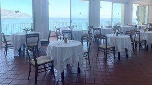 Un restaurante con mesas y sillas redondas, ventanas amplias con vista al mar y suelo de baldosas.