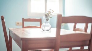 Uma mesa de jantar de madeira com um vaso de vidro transparente contendo uma planta, em frente a uma parede azul clara com uma janela ao fundo.