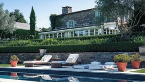 A pool area with lounge chairs and potted plants, surrounded by a stone wall and a large house in the background. - Villa Los Magnolios "Paderne" (Paderne)