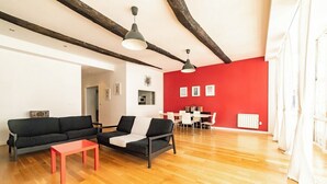 Un salon moderne avec un mur d’accent rouge, des poutres en bois, un canapé, une table basse et un coin repas avec des chaises blanches.