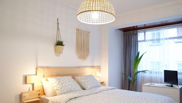 Ein Schlafzimmer mit Holzkopfteil, einem Bett mit Blumenbettwäsche, einem Nachttisch mit Lampe, einem hängenden Wandbehang aus Makramee und einem großen Fenster mit Vorhängen.