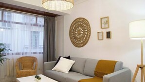 Ein modernes Wohnzimmer mit einem grauen Sofa, einem weißen Couchtisch und einem Korbstuhl. An der Wand befindet sich ein großes Dekorationskunstwerk, und es steht eine Stehlampe im Raum.