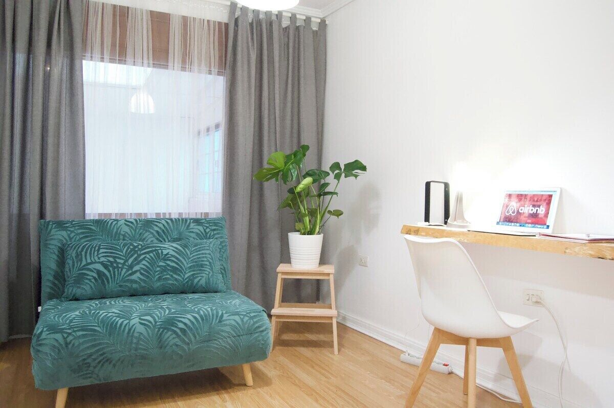 Una habitació amb un sofà color turquesa, un tamboret de fusta, una cadira blanca, una planta en test i un escriptori amb un ordinador portàtil i un cartell d'Airbnb.
