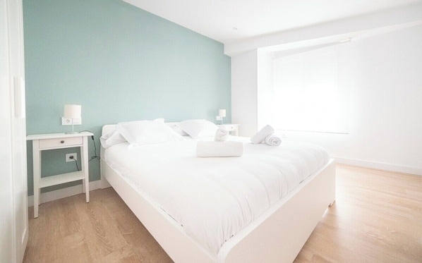 A modern bedroom with a large bed, white bedding, a nightstand with a lamp, and a window providing natural light.