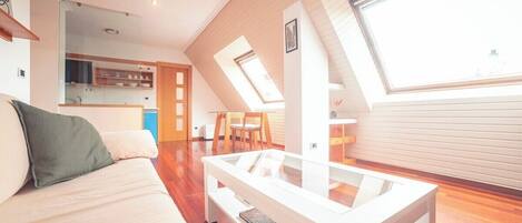 A modern loft apartment with a loft bed, a glass coffee table, and a kitchen area visible through an open door.